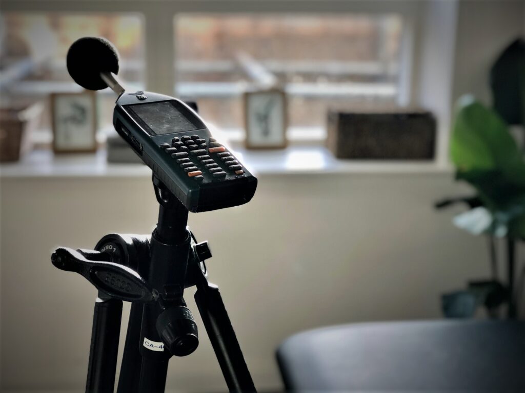 Close-up of a handheld device with a screen and keypad mounted on a tripod, with a foam-covered microphone above it in a home office setting.